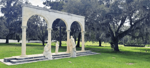 A serene outdoor scene featuring white stone sculptures under arches, surrounded by lush greenery and trees.