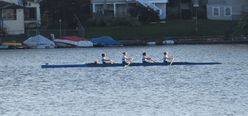 Four rowers in a blue boat glide across a calm lake, with houses and boats visible in the background.