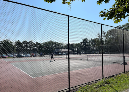 A tennis court surrounded by a chain-link fence, with a player practicing under a clear blue sky.