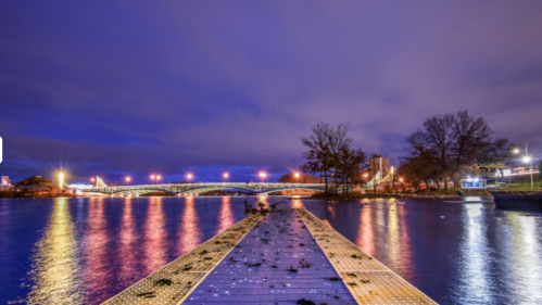 A serene river scene at dusk, featuring a wooden dock leading to a bridge illuminated by city lights.