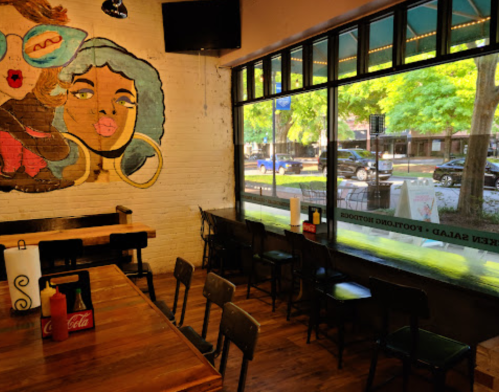 A cozy restaurant interior with a mural of two women, wooden tables, and large windows overlooking a tree-lined street.