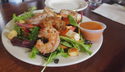 A plate of salad topped with seasoned shrimp, tomatoes, croutons, and a small cup of dressing on the side.