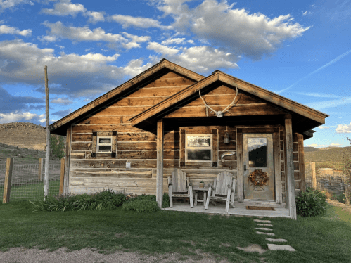A rustic wooden cabin with a porch, surrounded by grass and mountains under a blue sky with clouds.