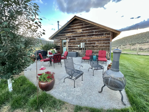 A cozy outdoor seating area with chairs and a table in front of a rustic wooden cabin, surrounded by greenery.