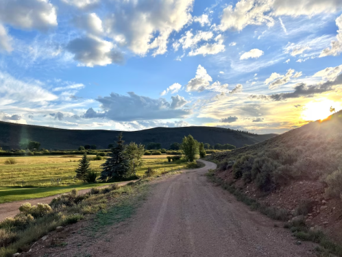 A winding dirt road leads through a lush green landscape under a colorful sky at sunset.