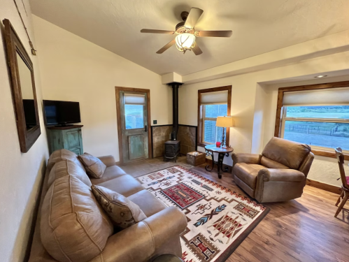 Cozy living room with a sofa, armchair, TV, and a view of the outdoors through large windows. Rustic decor and warm lighting.