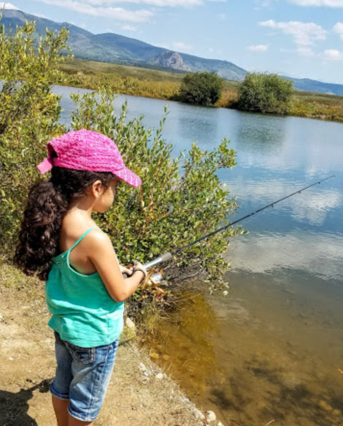 A child in a pink hat fishes by a calm lake surrounded by greenery and mountains under a blue sky.