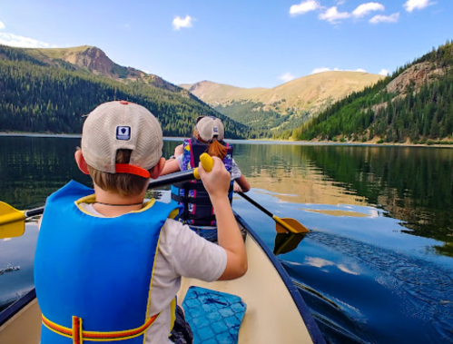 Two children in life jackets paddle a canoe on a calm lake surrounded by mountains and trees under a blue sky.