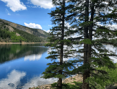 A serene lake surrounded by trees, reflecting blue skies and mountains in the background.