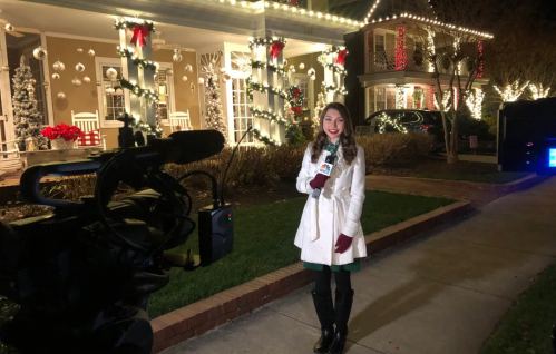 A reporter in a white coat stands outside a beautifully decorated house with holiday lights and ornaments.