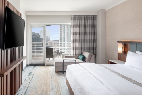 Modern hotel room featuring a king bed, seating area, and balcony with city views. Bright and airy decor.