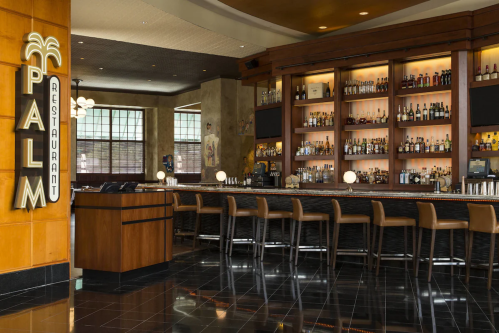 A stylish bar area with a wooden counter, high stools, and shelves filled with various bottles in a restaurant setting.