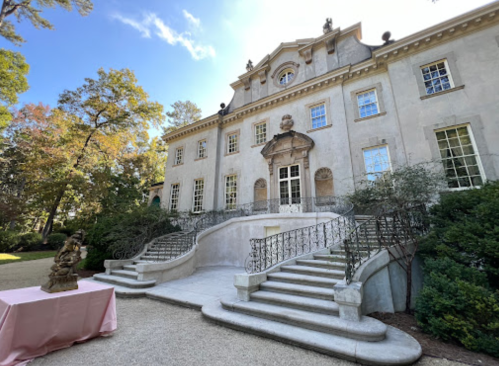 A grand mansion with a staircase, ornate facade, and lush greenery surrounding it on a sunny day.