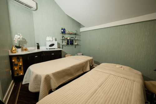 A serene spa room with two massage tables, soft lighting, and shelves of spa products against a green wall.