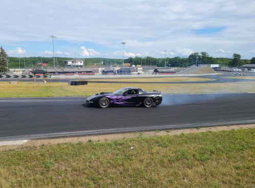 A black sports car with purple flames drifts on a racetrack, leaving tire smoke behind.