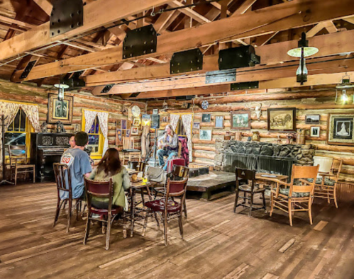 A cozy log cabin interior with wooden beams, a small stage, and people seated at tables enjoying the atmosphere.