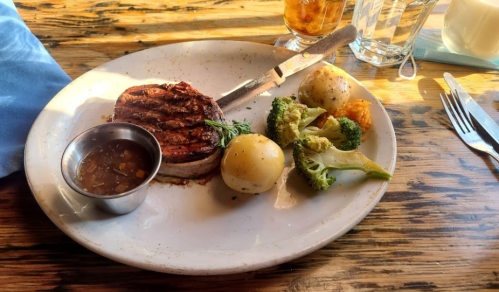 A plate with sliced meat, a small bowl of sauce, broccoli, potatoes, and a biscuit on a wooden table.