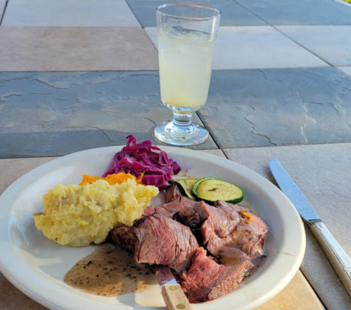 A plate with sliced beef, mashed potatoes, red cabbage, and zucchini, alongside a glass of lemonade on a table.