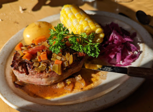 A plate with a grilled steak topped with vegetables, corn on the cob, a potato, and purple cabbage, garnished with parsley.