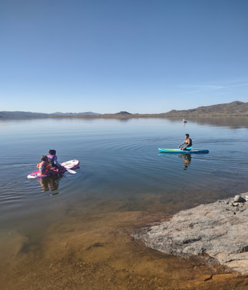 Two people paddleboarding on a calm lake, with mountains in the background and clear blue skies.