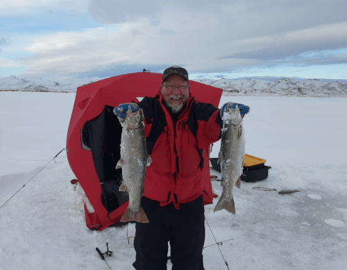 A smiling man in winter gear holds two large fish in a snowy landscape, with a red ice fishing shelter in the background.