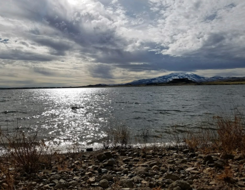 A serene lake scene with shimmering water, rocky shore, and mountains under a cloudy sky.
