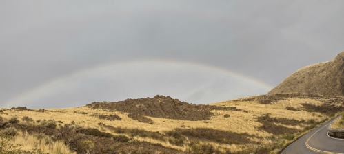A scenic view of a rainbow arching over rocky hills and dry grasslands, with a winding road in the foreground.