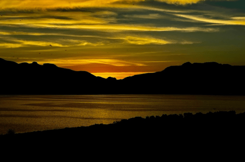 Silhouette of mountains against a vibrant sunset over a calm lake, with colorful clouds in the sky.