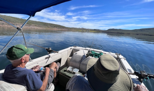 Two people sit in a boat on calm water, surrounded by hills and a clear blue sky, with fishing gear visible.