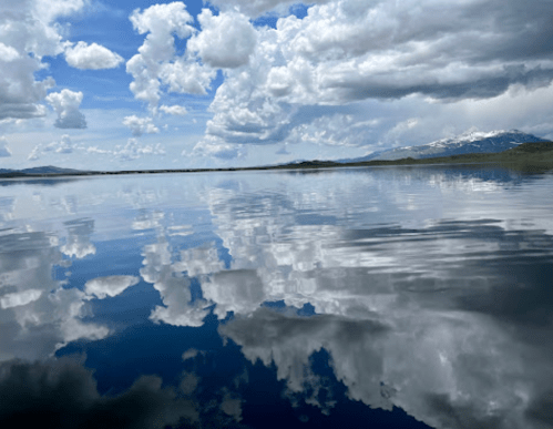 A serene lake reflecting fluffy clouds and mountains under a blue sky, creating a tranquil and picturesque scene.