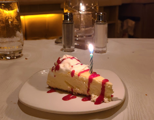 A slice of cheesecake topped with whipped cream and raspberry sauce, featuring a lit birthday candle on a white plate.