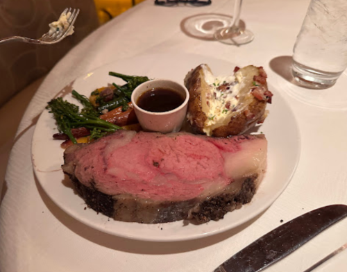 A plate featuring a large slice of prime rib, roasted vegetables, a baked potato, and a small cup of sauce.