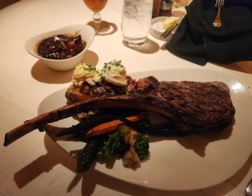 A large grilled ribeye steak with mashed potatoes and mixed vegetables on a white plate, served with a drink in the background.