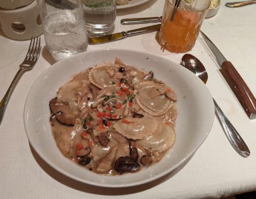 A bowl of ravioli in a creamy mushroom sauce, garnished with herbs and diced vegetables, on a dining table.