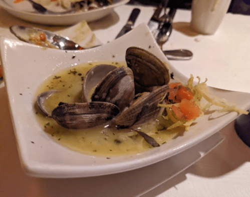 A white bowl filled with clams in a broth, garnished with herbs and diced tomatoes, on a table setting.