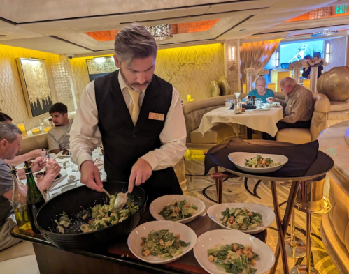 A waiter serves salad at a fine dining restaurant, with guests dining in the background. Elegant decor surrounds them.