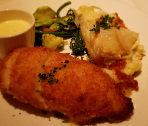 A plate featuring breaded fish, creamy mashed potatoes, sautéed vegetables, and a small cup of sauce.