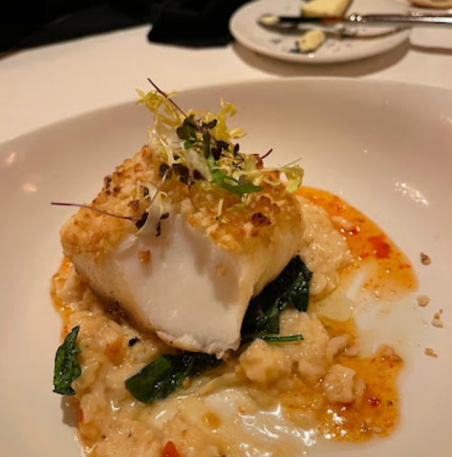 A beautifully plated dish featuring a piece of fish on a bed of creamy risotto with greens and a drizzle of sauce.