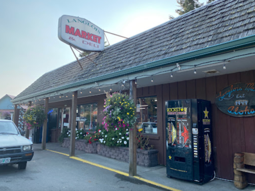 Exterior of Langlois Market & Deli, featuring a sign, flower baskets, and a vending machine outside.