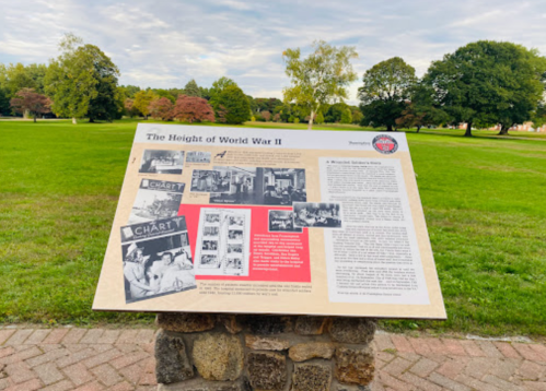 Informational sign about World War II, featuring images and text, set in a grassy park with trees in the background.