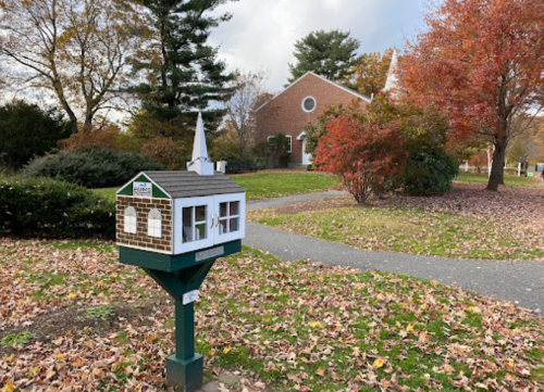 A whimsical mailbox shaped like a house, surrounded by autumn leaves and trees, with a church in the background.