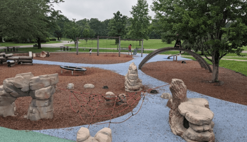 A playground with climbing structures, a sandbox, and grassy areas, surrounded by trees and picnic tables.