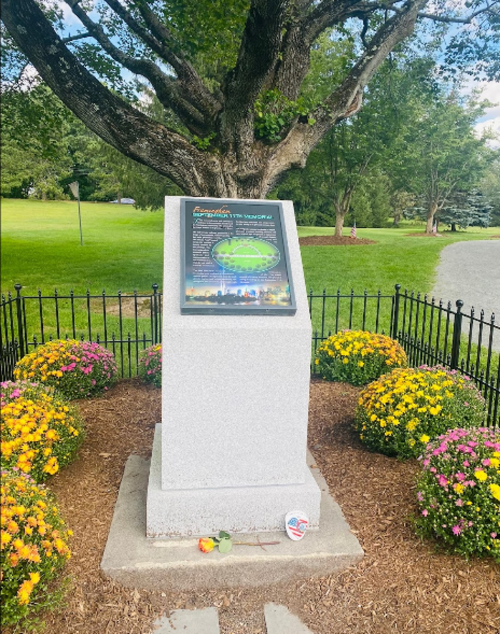 A stone monument with an informational plaque surrounded by colorful flowers and a tree in a park setting.