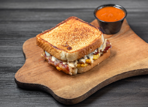 Grilled sandwich with melted cheese and bacon, served on a wooden board with a side of dipping sauce.