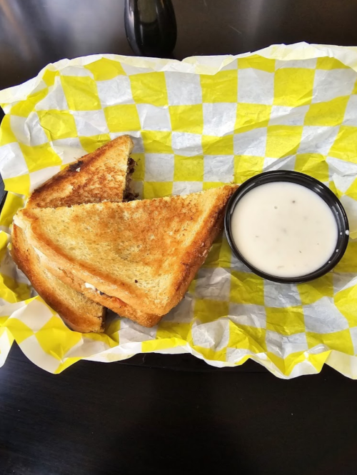 Grilled cheese sandwich on yellow checkered paper with a small cup of creamy dipping sauce.
