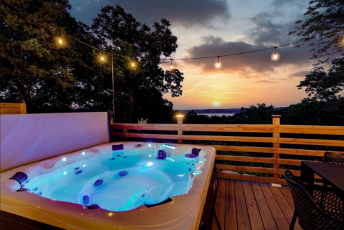 A hot tub on a wooden deck with string lights, overlooking a sunset and lush trees.