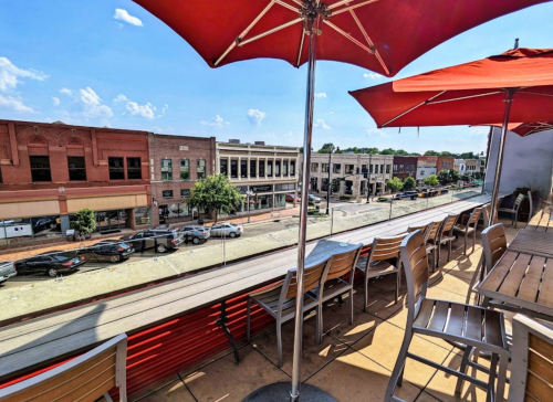 A rooftop patio with red umbrellas overlooks a busy street lined with shops and cars under a clear blue sky.