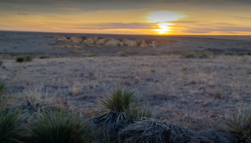 A serene sunset over a vast, dry landscape with sparse vegetation and distant cliffs.
