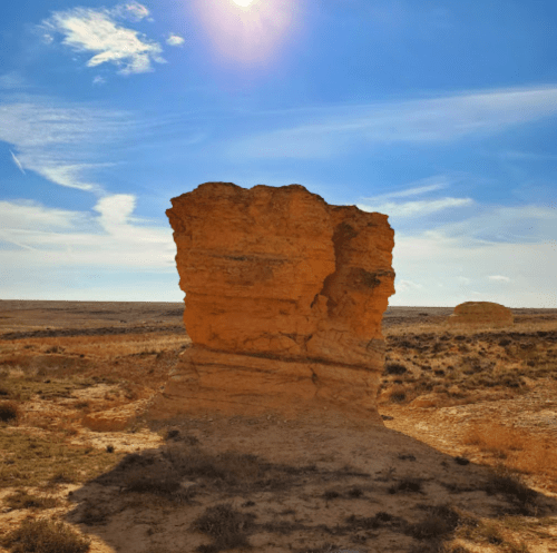 A tall, weathered rock formation stands against a clear blue sky, with the sun shining above in a desert landscape.