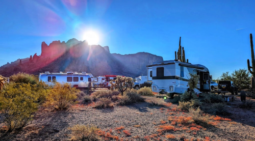 Sunrise over rugged mountains, RVs parked among desert vegetation, with cacti and a clear blue sky.